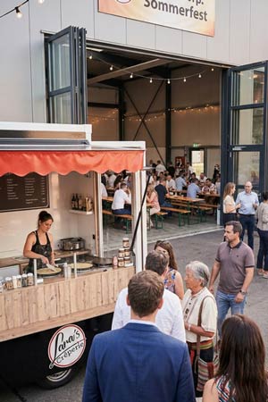 Vor einer großen offenen Halle, in der Gruppen an langen Tischen eine sommerliche Firmenfeier genießen, stehen die Menschen am Reicherts-Crepe-Imbisswagen an. Im Inneren des Trucks bereitet eine Frau frische Leckereien zu und präsentiert Mobiles Catering unter einem roten Baldachin.