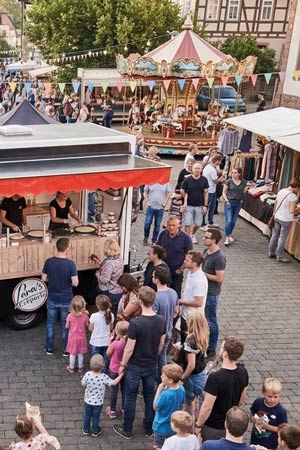 Ein lebhaftes Stadtfest unter freiem Himmel mit Menschen, die sich um einen mobilen Imbisswagen scharen, Kindern und Erwachsenen, die Schlange stehen, Marktständen und einem alten Karussell im Hintergrund. Die Atmosphäre ist festlich und fröhlich.