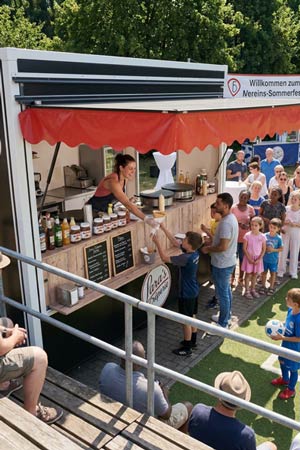 Bei einer Sommerveranstaltung eines Vereins im Freien serviert eine Frau einem Jungen Crepes aus einem Foodtruck von Reicherts Mobiles Catering, während Kinder und Erwachsene unter sonnigem Himmel und mit Bäumen im Hintergrund in der Schlange warten.