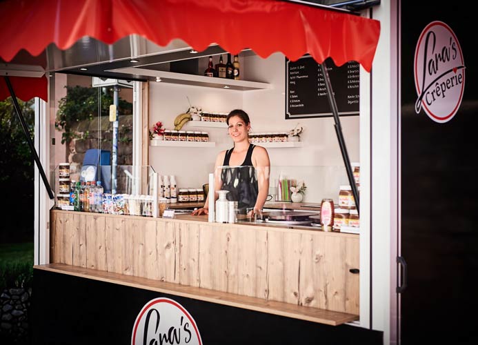 Eine Frau, Lara Reicherts, steht lächelnd und mit schwarzer Schürze hinter der Theke eines hölzernen Imbisswagens mit der Aufschrift Lanas Crêperie. Im Hintergrund sind Regale mit Zutaten und eine Menükarte zu sehen, die ideal für das Catering auf Eventmobilen sind.