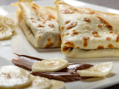 Zwei gefaltete Crêpes auf einem Teller, serviert mit Bananenscheiben und einem Schokoladen-Haselnuss-Aufstrich, wahrscheinlich Nutella, als Beilage.