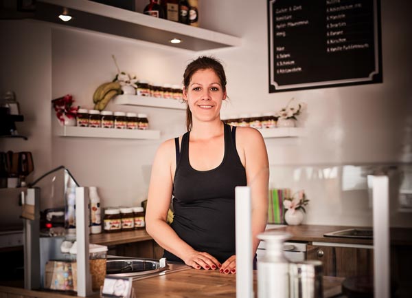 Lara Reichert, in einem schwarzen Tank-Top, steht lächelnd hinter dem Tresen eines kleinen Ladens oder Cafés. Im Hintergrund sind Regale mit Gläsern, Bananen und eine Tafel mit der Speisekarte zu sehen.