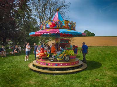 Ein buntes, kleines Karussell, auf dem Kinder reiten, steht draußen auf der Wiese beim Reicherts Maifest. Mehrere Erwachsene und Kinder stehen in der Nähe unter einem klaren blauen Himmel, mit Feldern und Bäumen im Hintergrund.